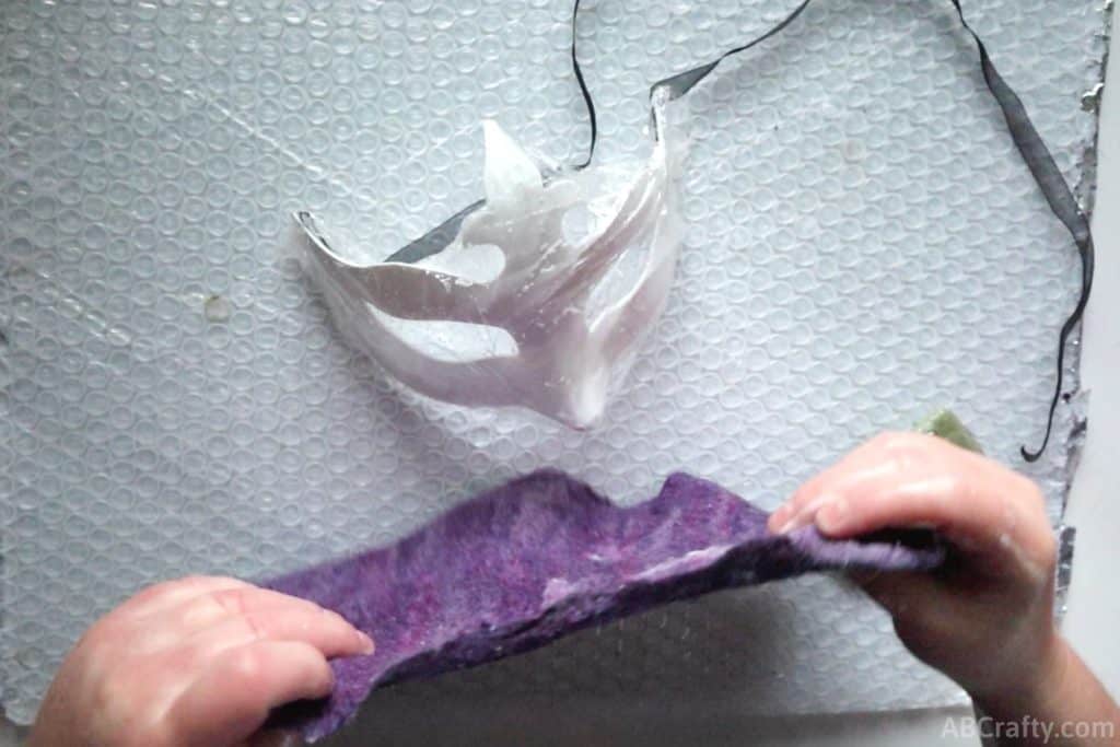placing a partially felted purple piece of wool fabric over a plastic white Venetian mask covered in plastic wrap