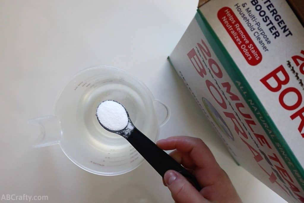 holding a teaspoon of borax powder over a clear measuring cup with water