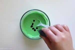 adding drops of green food coloring to a bowl with green liquid with a plastic spoon in it