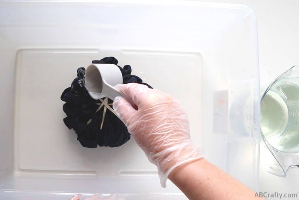 pouring bleach mixture from measuring cup onto a bundled up blue shirt, sitting a plastic tub