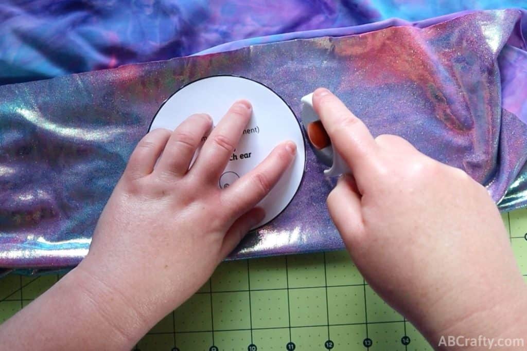 using a rotary cutter to cut around the ab crafty mickey ear template on blue and purple iridescent fabric