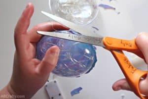 using fabric scissors to cut around blue and purple iridescent fabric glued to a clear plastic ornament