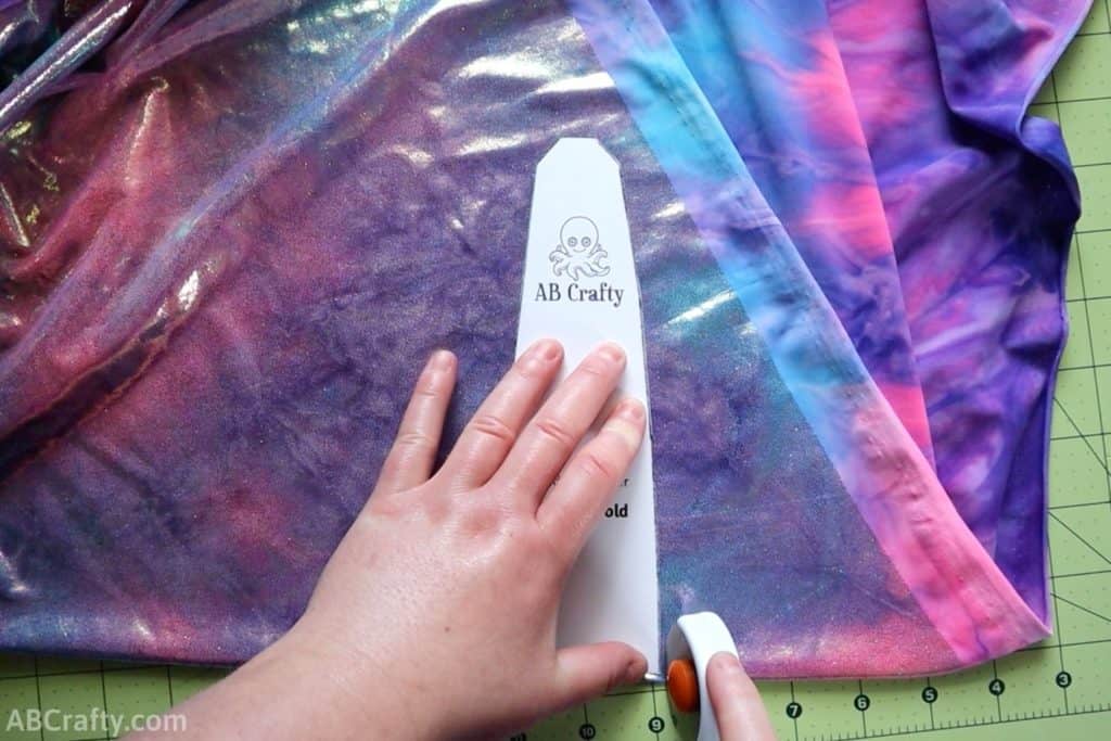 using a rotary cutter to cut purple and blue iridescent fabric around the ab crafty headband template