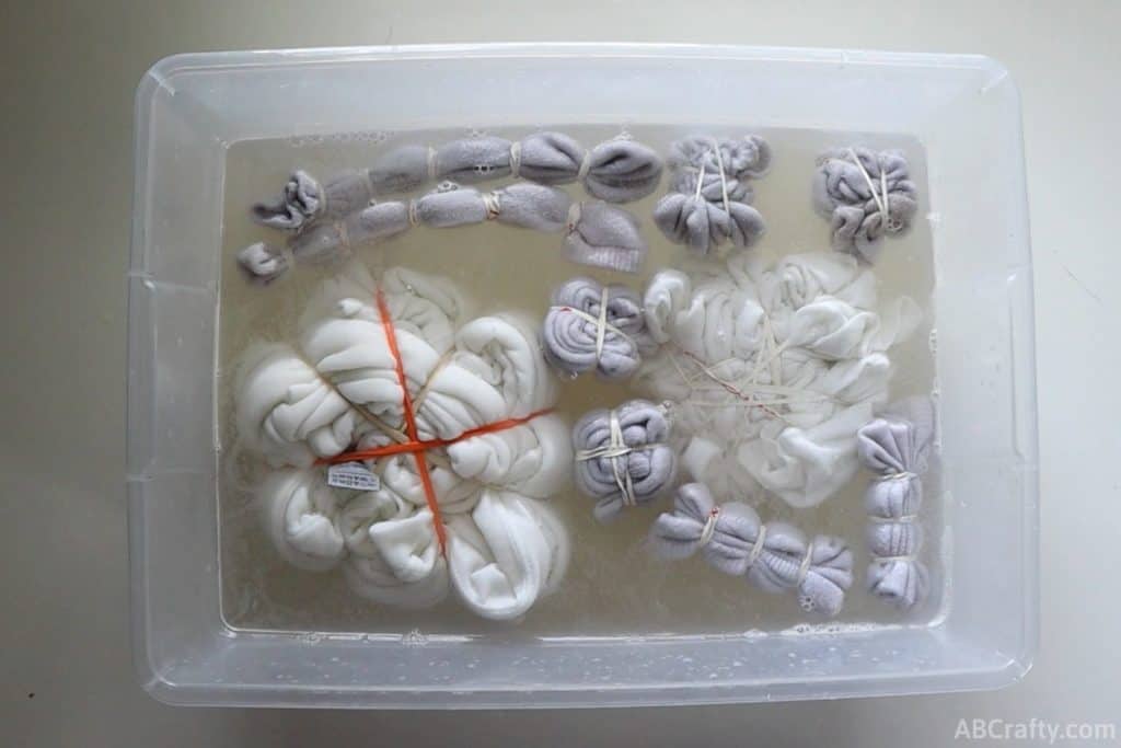 multiple pieces of white clothing wrapped in rubber bands and soaking in water and soda ash in a plastic bin