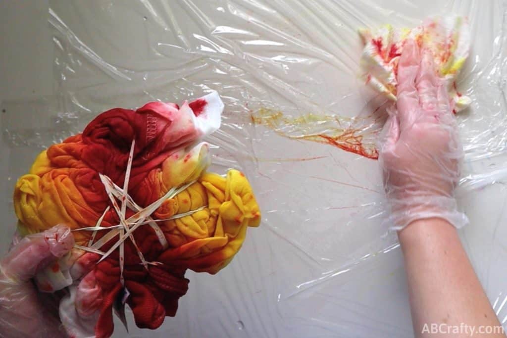 using a paper towel to wipe dye off of plastic wrap on a table while holding a partially tie dyed shirt