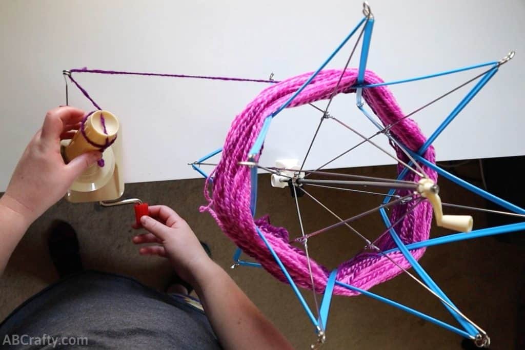 turning the handle of a yarn ball winder to begin winding pink yarn off a yarn swift