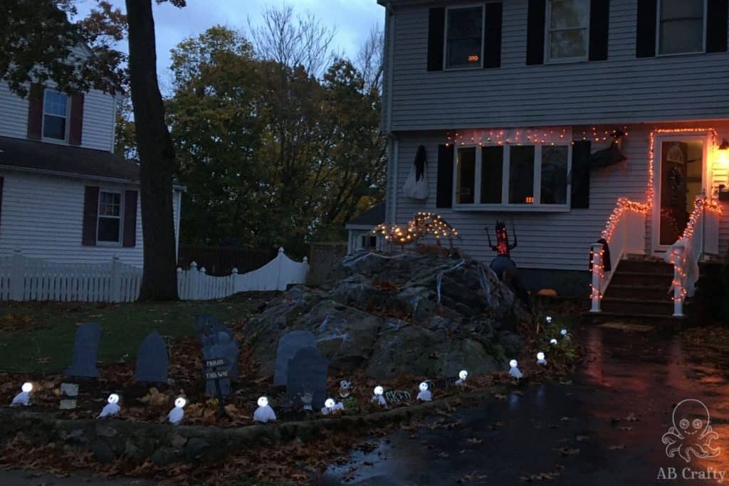 halloween decorations in front of a house including ghost pathway lights, a light up animatronic spider, gravestones, and halloween lights hanging from the house