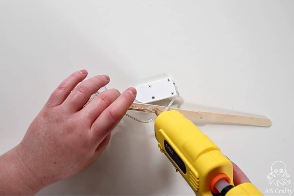 using a glue gun to add glue to a popsicle stick with fairy lights hanging off