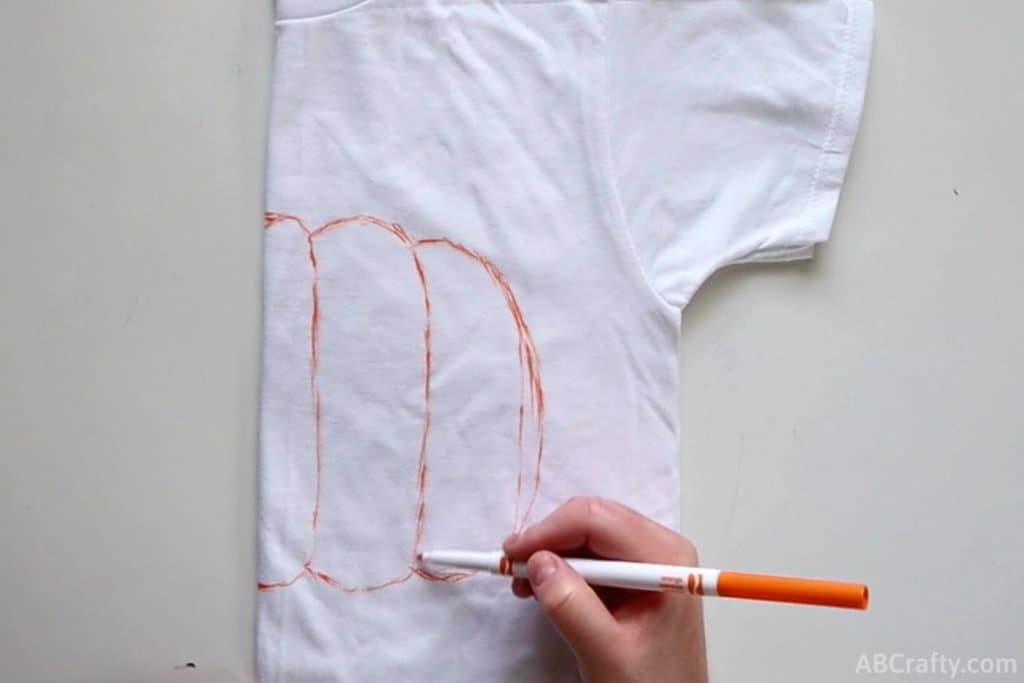 using an orange washable marker to draw the inside lines of a pumpkin on a folded white shirt