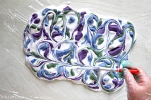 using the back of a pen to draw a line through swirls in shaving cream with drops of blue, purple, and green dyed shaving cream