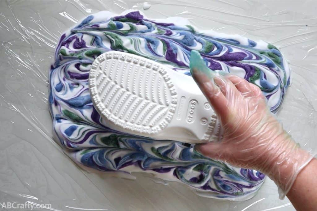 putting a white croc into a rectangle of shaving cream with blue, purple, and green shaving cream swirled throughout