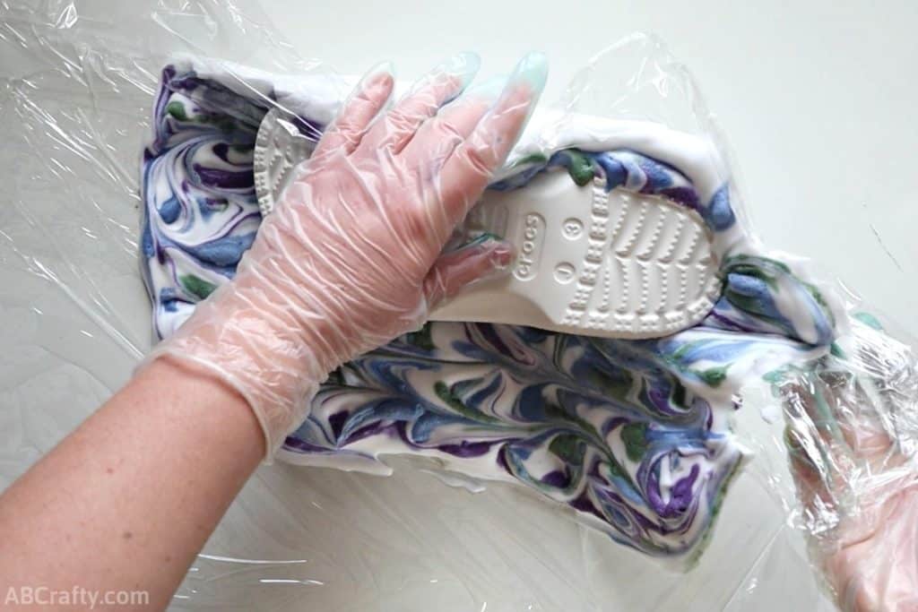 pulling up the plastic to the sides of a white croc with shaving cream with blue, purple, and green shaving cream swirled throughout