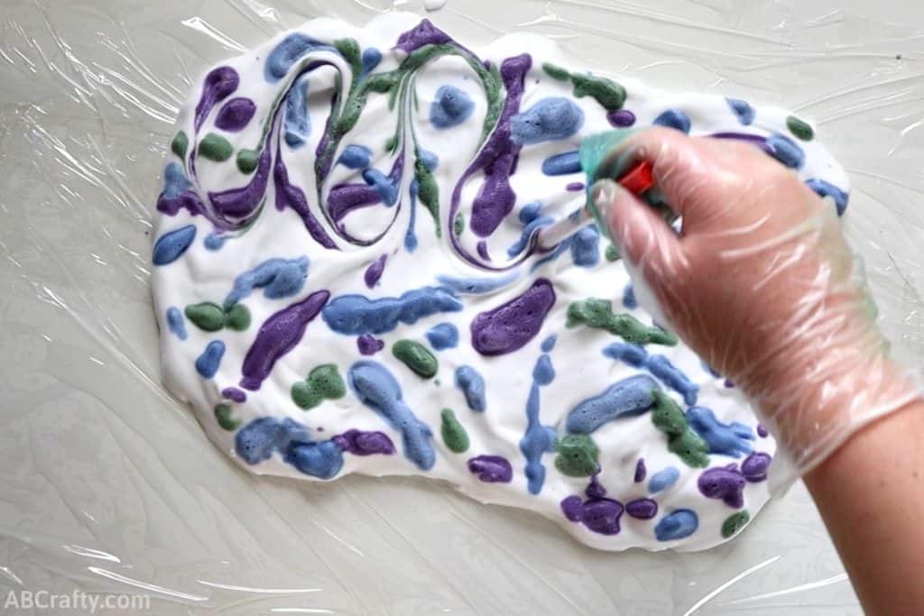 using the back of a pen to draw swirls in shaving cream with drops of blue, purple, and green dyed shaving cream