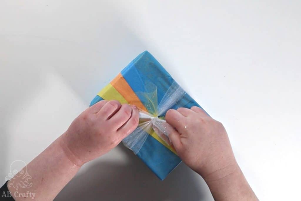 tying white tulle around a strip of yellow tulle to form a bow on a present