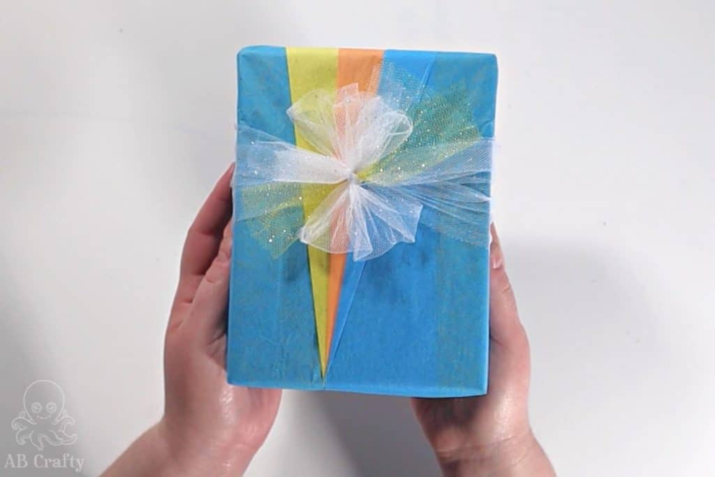 holding the finished origami style wrapped present with blue yellow and orange tissue paper forming triangles and a ribbon made of white tulle
