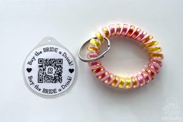 laminated and printed buy the bride a drink venmo qr code with hole punched at the top next to a pink, orange, and yellow keychain bracelet