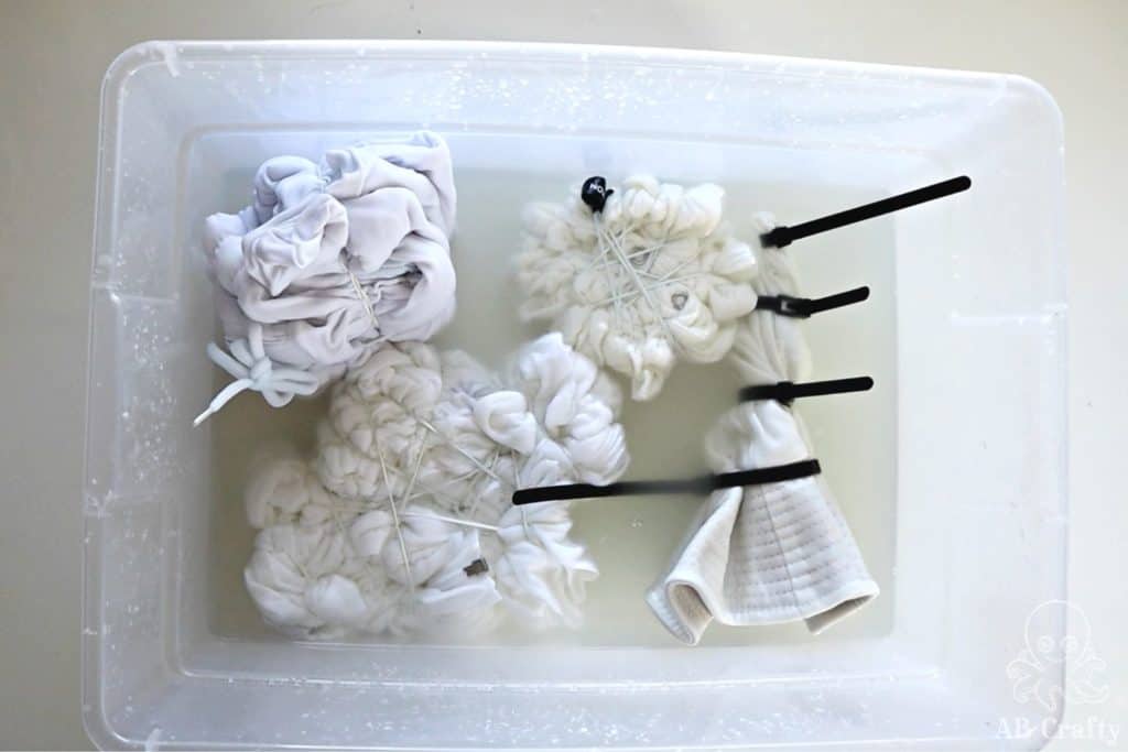 white cotton clothing twisted and tied with zip ties and rubber bands and soaking in a bucket of soda ash solution