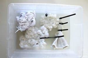 white cotton clothing twisted and tied with zip ties and rubber bands and soaking in a bucket of soda ash solution