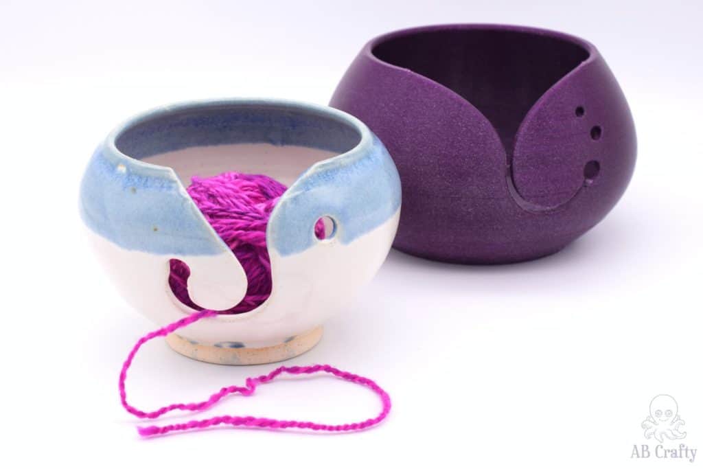 a blue and white ceramic yarn bowl with pink yarn inside and a purple glitter printed yarn bowl