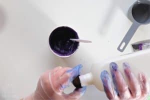 pouring acetone into the bottle cap next to a paper cup with shaving cream and purple dye