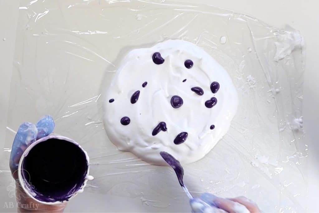 using a spoon to add drops of purple dye onto a circle of shaving cream on plastic wrap
