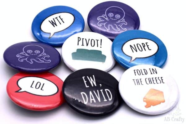 finished homemade buttons with the different sayings, including an "ew, david" button, a "pivot!" button with a couch, "fold in the cheese" button with a cheese block, and buttons with "wtf" "lol" and "nope" and the AB Crafty logo