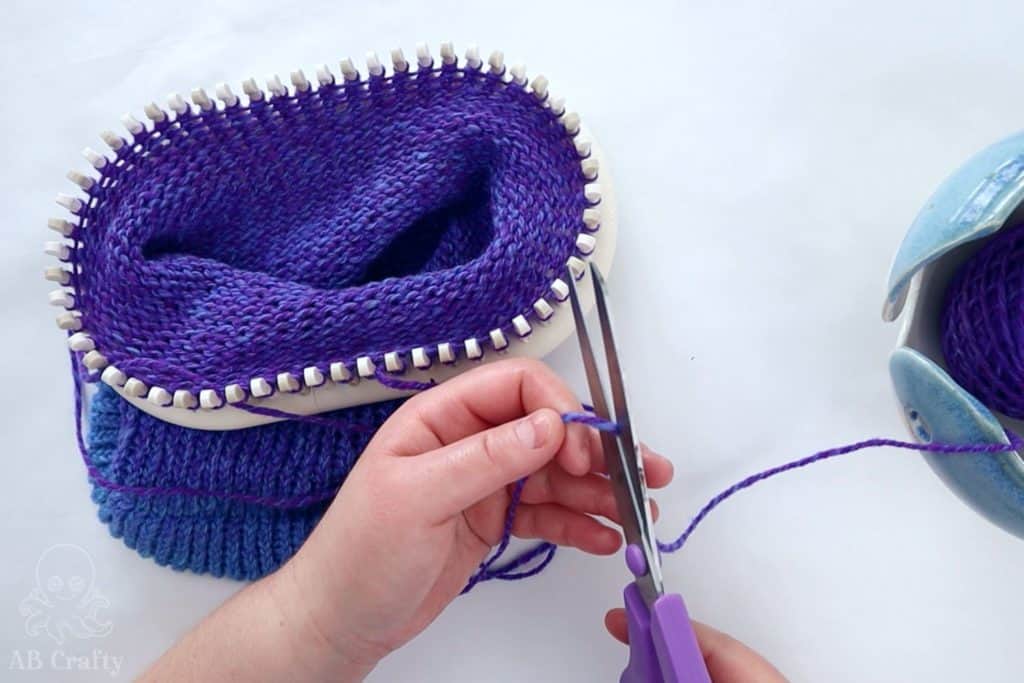 cutting the purple yarn with the knit project on the knitting loom behind it