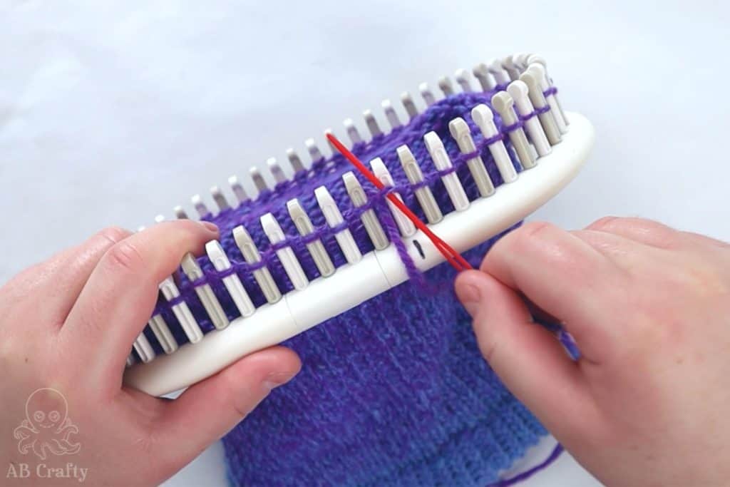 using a red plastic needle to go under the first peg of the knitting loom, under the first stitch