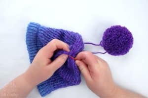 using a red plastic needle to sew the string of a pom pom into the top of the loom knitting hat