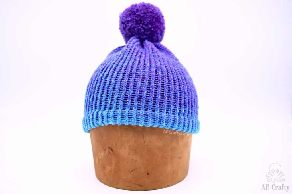 finished blue and purple loom knitting hat with a double brim and purple pom pom on a wooden hat block