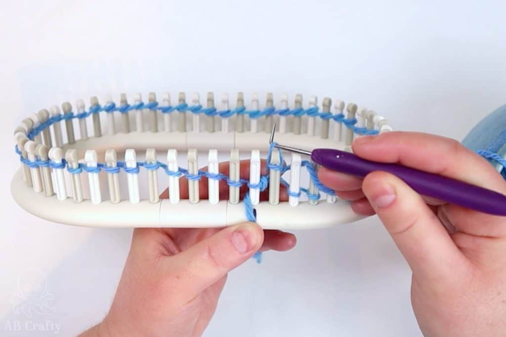 using a knitting loom hook to lift the stitch over the peg of the loom