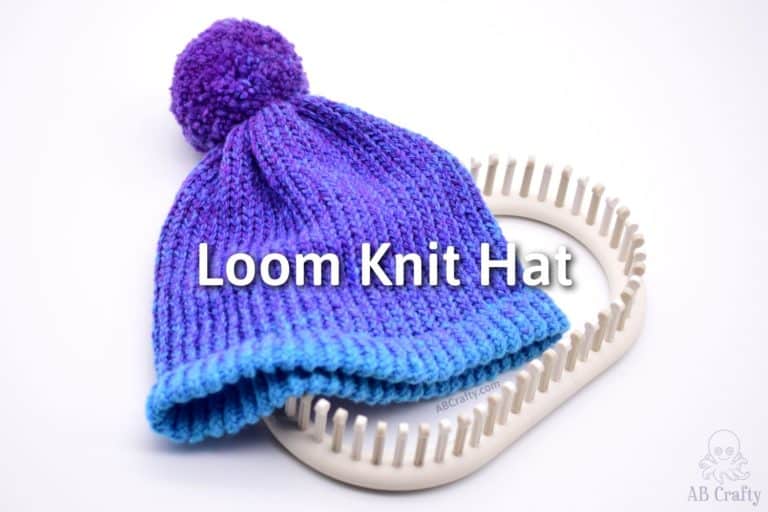 finished blue and purple knit beanie with a double brim and purple pom pom on top of the adjustable knitting loom with the title "loom knit hat"