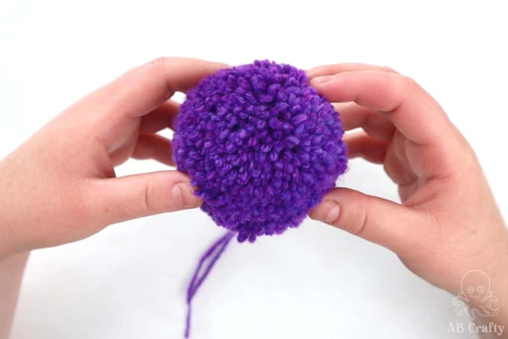 holding a finished purple pom pom with the tied ends hanging down