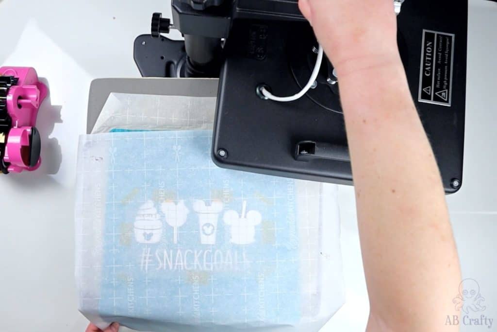 pushing the top of a swing heat press away, revealing the bottom plate with the pressed item, design, and parchment paper
