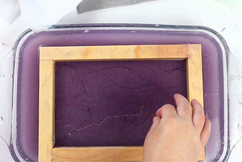placing threads of different colors inside the paper making frame over purple paper pulp